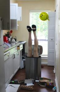 Person upside down in trash can, kitchen mess nearby.