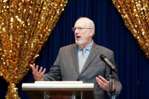 Man speaking at podium before gold decorative backdrop