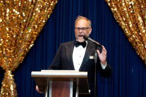 Man in tuxedo speaking at podium beneath gold decorations